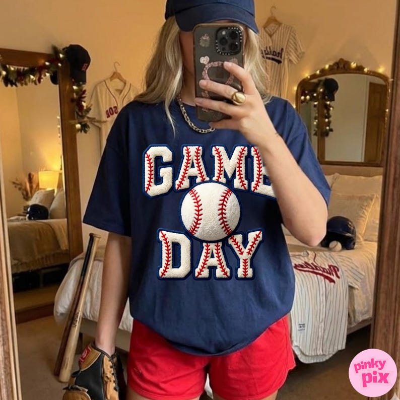 May include: Navy blue t-shirt with the words "GAME DAY" in white and red, featuring a baseball graphic. The person is wearing red shorts and a navy blue baseball cap. Baseball theme.