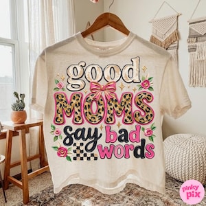 May include: Cream-colored t-shirt with the text "good MOMS say bad words" in a playful design. The word "MOMS" features a leopard print pattern, and the design includes pink accents and floral details.