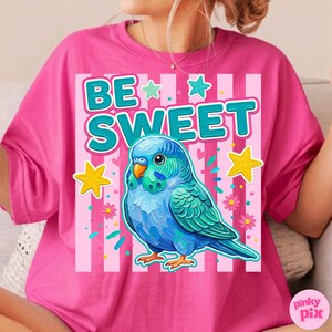 May include: A pink t-shirt featuring a blue parakeet illustration with the words "BE SWEET" in turquoise. The design includes stars, flowers, and vertical pink stripes. The shirt is a vibrant, eye-catching piece.