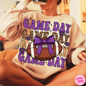 May include: A beige sweatshirt with a purple and brown football graphic. The graphic includes the words "GAME DAY" in purple, a football with a purple bow, and floral accents. The sweatshirt has long sleeves.