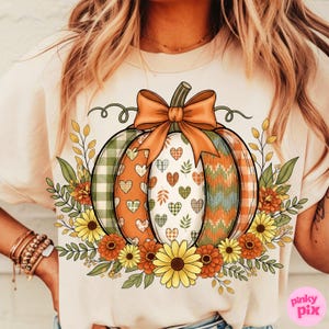 May include: Cream-colored t-shirt featuring a colorful pumpkin design. The pumpkin is decorated with plaid, hearts, and chevron patterns, topped with an orange bow and surrounded by fall foliage and flowers. The design is perfect for autumn.