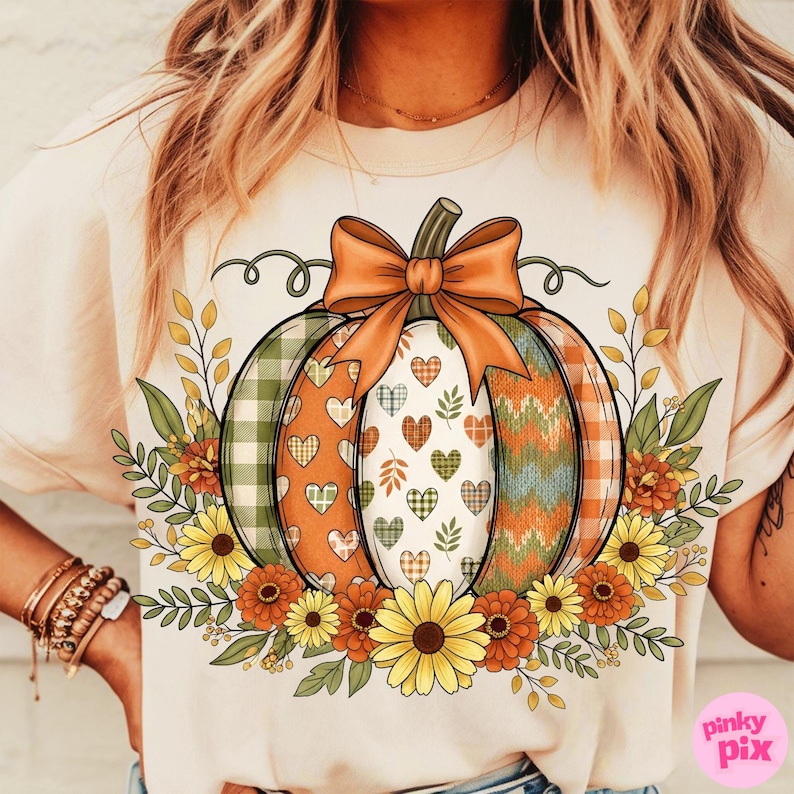 May include: Cream-colored t-shirt featuring a colorful pumpkin design. The pumpkin is decorated with plaid, hearts, and chevron patterns, topped with an orange bow and surrounded by fall foliage and flowers. The design is perfect for autumn.