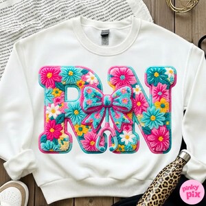 May include: White sweatshirt featuring the letters "RN" in a floral design. The letters are filled with pink, blue, yellow, and white flowers. A bow and stethoscope are in the center. The sweatshirt is a crew neck.