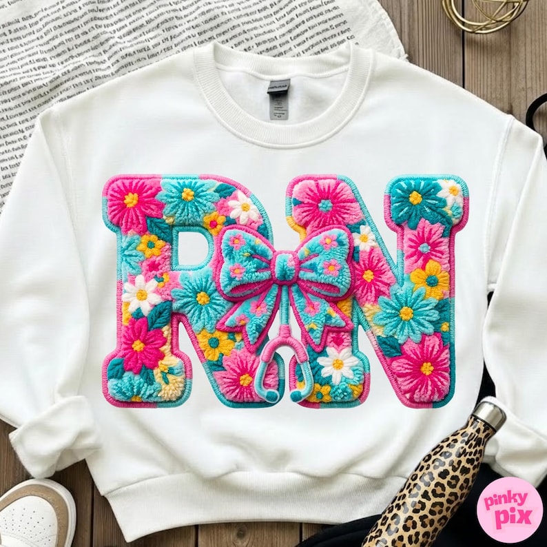 May include: White sweatshirt featuring the letters "RN" in a floral design. The letters are filled with pink, blue, yellow, and white flowers. A bow and stethoscope are in the center. The sweatshirt is a crew neck.