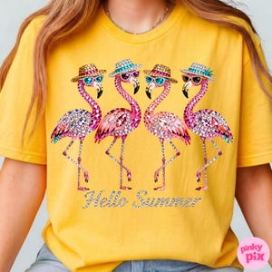May include: Yellow t-shirt featuring four pink flamingo illustrations. Each flamingo wears sunglasses and a hat, adorned with sparkling gems. The phrase "Hello Summer" is written in silver glitter. The shirt is a casual summer wear.
