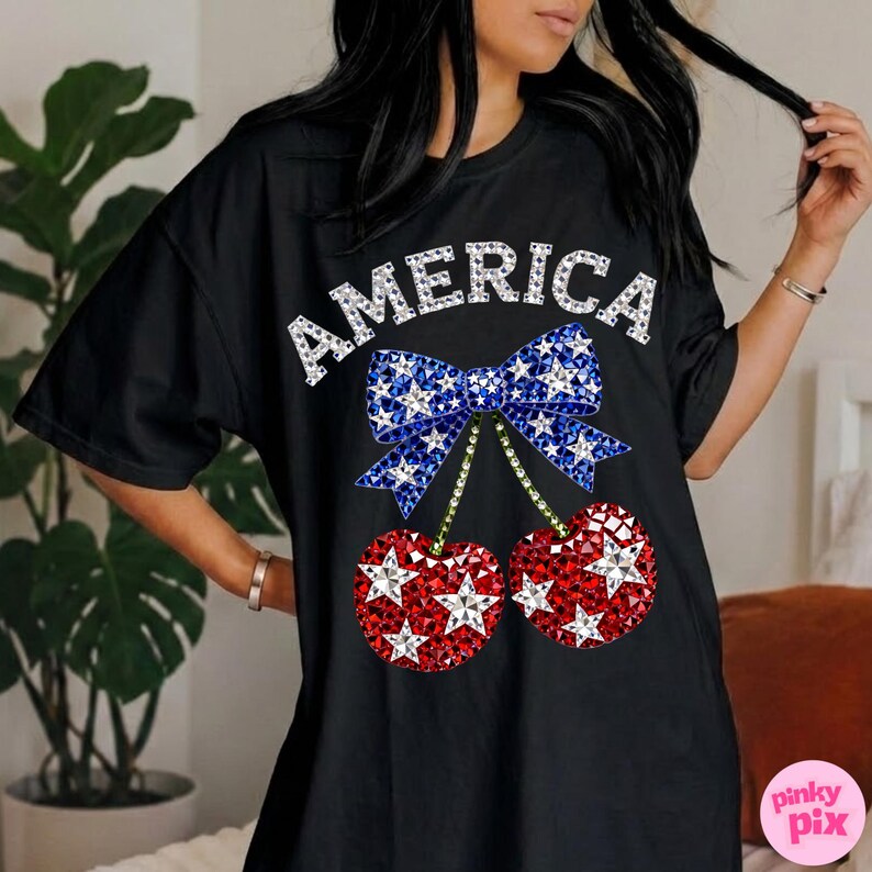 May include: Black t-shirt with the word "AMERICA" in white rhinestone lettering. The shirt features a blue bow with white stars and two red cherries with white stars, all in a rhinestone design.