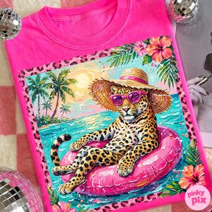 May include: Bright pink t-shirt with a colourful illustration of a leopard wearing sunglasses and a straw hat, relaxing on a pink inflatable ring in the sea. The design includes palm trees, flowers, and a tropical beach scene.
