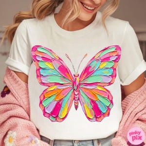 May include: White t-shirt featuring a vibrant, colorful butterfly design. The butterfly's wings are painted with thick brushstrokes in shades of pink, green, yellow, and orange. The shirt has short sleeves and a crew neck. The brand name "pinky pix" is in the bottom right corner.