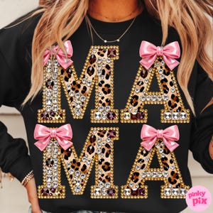 May include: Black sweatshirt featuring the word "MAMA" in large, embellished letters. The letters have a gold outline and leopard print filling. Pink bows are attached to the top of each letter.