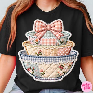 Puede incluir: Camiseta negra con un gr&aacute;fico de tres cuencos decorativos apilados. Los cuencos tienen un dise&ntilde;o de patchwork con estampados florales y de cuadros, y un lazo rosa. El dise&ntilde;o tiene una apariencia acolchada con un borde festoneado.