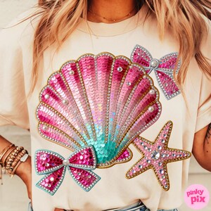 May include: A light beige t-shirt featuring a large, sequined seashell design in shades of pink and teal, adorned with pearls and rhinestones. Two sequined bows and a starfish are also featured. The design has a gold trim.