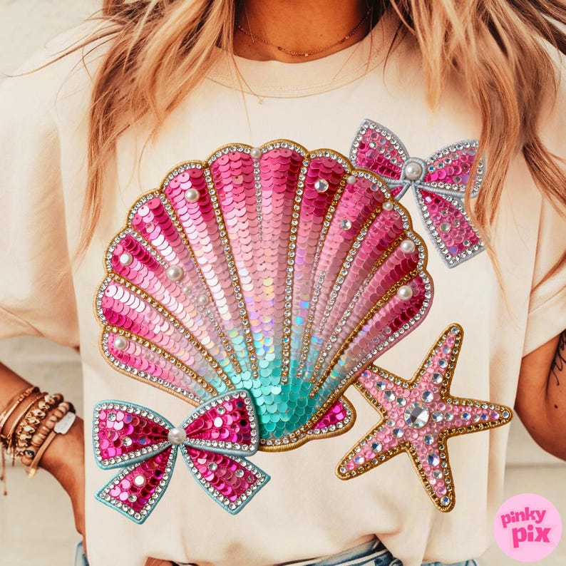 May include: A light beige t-shirt featuring a large, sequined seashell design in shades of pink and teal, adorned with pearls and rhinestones. Two sequined bows and a starfish are also featured. The design has a gold trim.