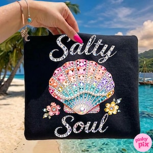 Puede incluir: Bolso tote negro con las palabras "Salty Soul" en una fuente brillante. El bolso presenta un diseño de concha marina colorido con detalles de cristal y adornos florales. El fondo muestra una escena de playa.