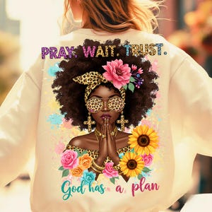 May include: A cream-colored sweatshirt with a graphic print of a woman with brown hair wearing leopard print sunglasses and a headband. The graphic also includes flowers, sunflowers, and the text "Pray. Wait. Trust. God has a plan."