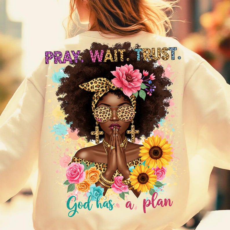 May include: A cream-colored sweatshirt with a graphic print of a woman with brown hair wearing leopard print sunglasses and a headband. The graphic also includes flowers, sunflowers, and the text "Pray. Wait. Trust. God has a plan."
