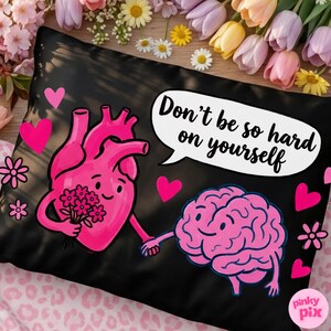 May include: Black pillowcase with a pink heart and brain illustration holding hands. The heart holds flowers. Text in a speech bubble reads, "Don't be so hard on yourself." Pink hearts and flowers decorate the pillowcase.
