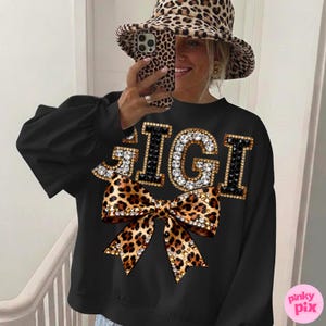 May include: Black sweatshirt with the word "GIGI" in rhinestone-studded letters and a leopard print bow. The sweatshirt has long sleeves. A matching leopard print hat is also visible.