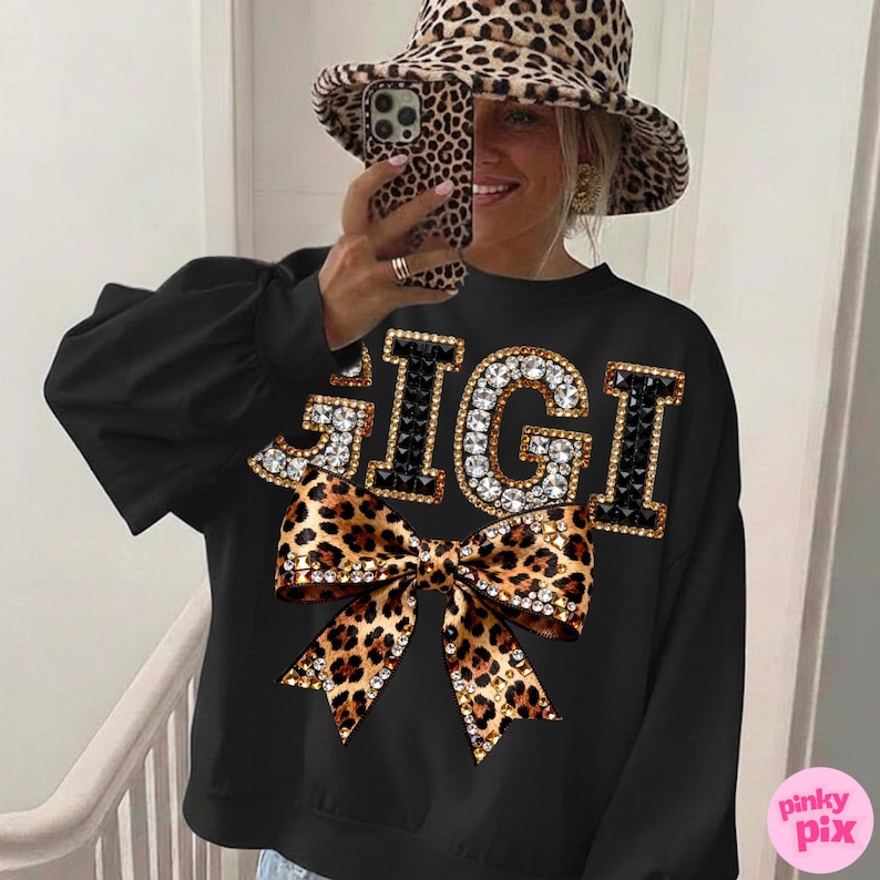 May include: Black sweatshirt with the word "GIGI" in rhinestone-studded letters and a leopard print bow. The sweatshirt has long sleeves. A matching leopard print hat is also visible.