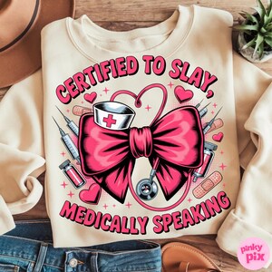 May include: Cream-colored sweatshirt with a pink graphic. The graphic features a large pink bow, a nurse's hat with a red cross, syringes, hearts, and the text "CERTIFIED TO SLAY, MEDICALLY SPEAKING" in pink.