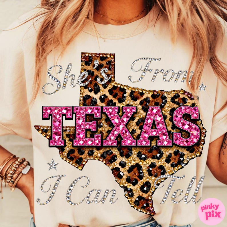 May include: A beige t-shirt featuring a leopard print Texas outline with the word "TEXAS" in pink, rhinestone-studded letters. The text "She's From" and "I Can Tell" are written in rhinestone lettering, with small star accents.