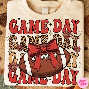 May include: Cream-colored sweatshirt with the words "GAME DAY" in red, leopard print, and tan lettering. A football with a red bow is in the center. Floral accents and stars complete the design.