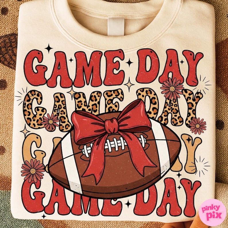 May include: Cream-colored sweatshirt with the words "GAME DAY" in red, leopard print, and tan lettering. A football with a red bow is in the center. Floral accents and stars complete the design.