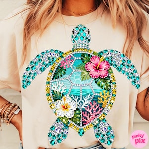 May include: A light beige t-shirt featuring a large, bejeweled sea turtle design. The turtle has turquoise gem-studded flippers and head, with a shell design of tropical flowers, coral, and ocean imagery. The shirt has a crew neck.