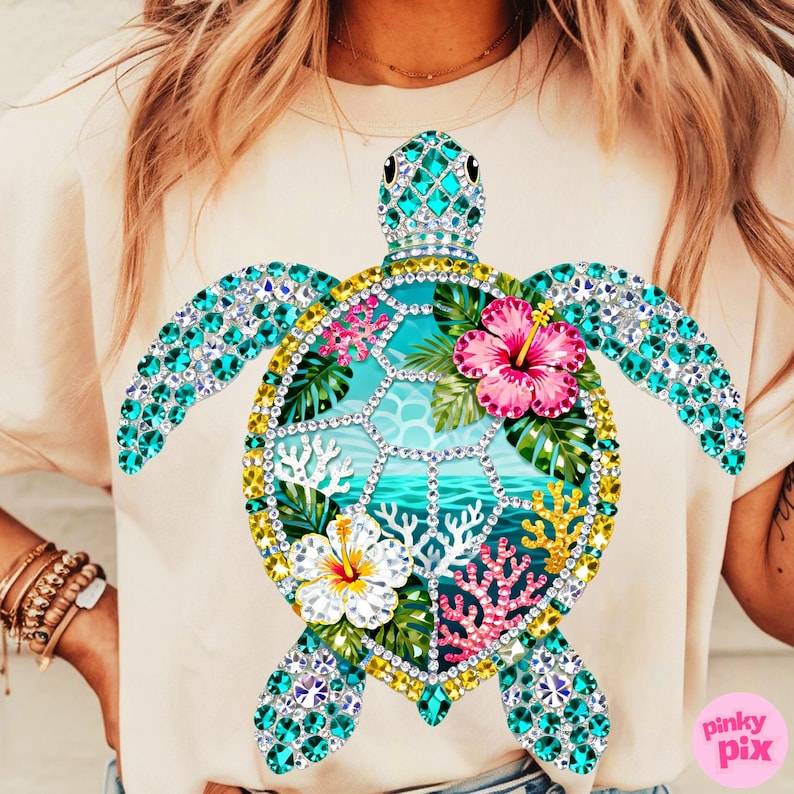 May include: A light beige t-shirt featuring a large, bejeweled sea turtle design. The turtle has turquoise gem-studded flippers and head, with a shell design of tropical flowers, coral, and ocean imagery. The shirt has a crew neck.