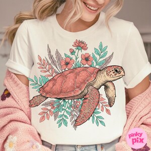 May include: White t-shirt with a pink and green floral design featuring a sea turtle. The turtle is outlined in black and has a pink shell.