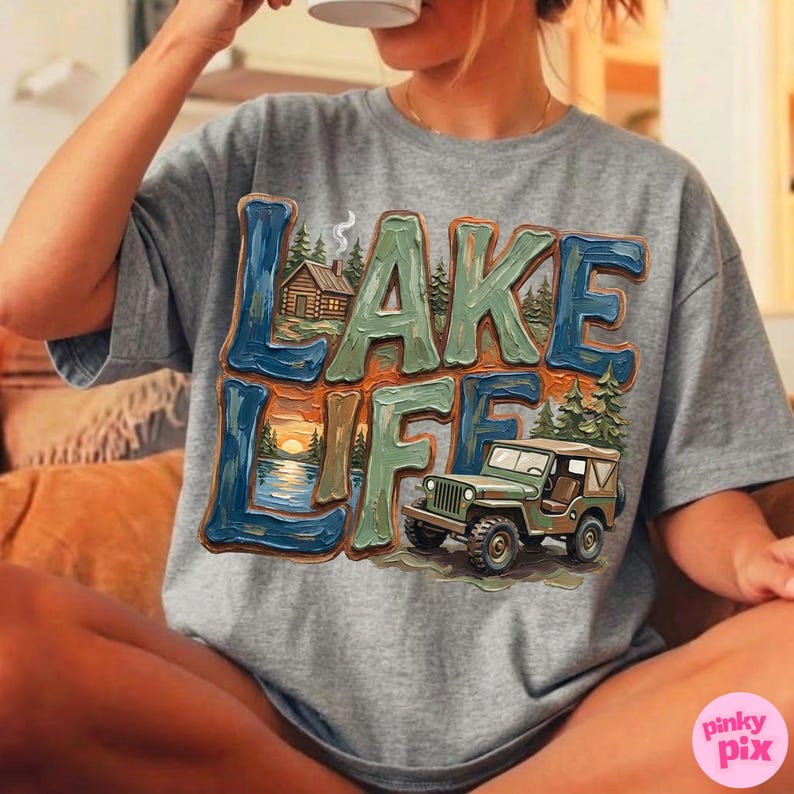 May include: A gray t-shirt featuring the words "LAKE LIFE" in a colorful, textured design. The graphic includes a cabin, a sunset over a lake, and a green jeep. The shirt is a comfortable, casual style.