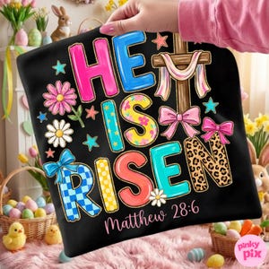 Könnte beinhalten: Schwarzes Kissen zum Thema Ostern mit dem Schriftzug "HE IS RISEN" in bunten, gemusterten Buchstaben. Ein Holzkreuz mit drapiertem Stoff und Schleifen ist abgebildet. Der Text "Matthew 28:6" ist ebenfalls enthalten. Dekorative Blumen und Sterne umgeben den Text.