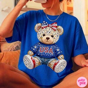 May include: A royal blue t-shirt featuring a bejeweled teddy bear design. The bear wears a blue sweater with "USA" in red letters, denim shorts, and red, white, and blue accents. A red, white, and blue bow adorns the bear's head.