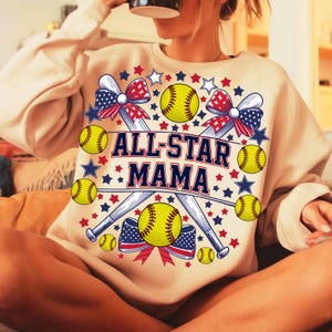 May include: Beige sweatshirt with the text "ALL-STAR MAMA" in navy blue. The design includes yellow softballs, crossed baseball bats, red, white, and blue bows, and stars. A sporty and patriotic theme.