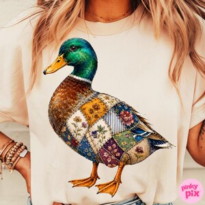 May include: A light beige t-shirt featuring a graphic of a mallard duck with a green head and yellow beak. The duck's body is composed of a patchwork design made of various floral and striped fabric patterns in shades of blue, yellow, and purple.
