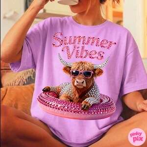 May include: A light purple t-shirt with the text "Summer Vibes" in pink rhinestone lettering. The shirt features a brown cow wearing sunglasses and a rhinestone-studded floatie. The cow is also adorned with rhinestones.