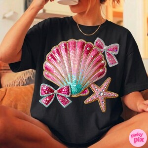 May include: Black t-shirt featuring a sequined seashell design in pink, teal, and gold. The design includes a starfish and two bows, all embellished with rhinestones. The shirt has a crew neck and short sleeves.
