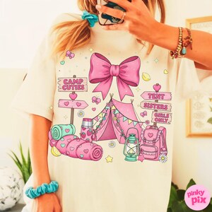 May include: Cream t-shirt with a pink camping design. The design includes a tent, bow, and signs reading "Camp Cuties" and "Tent Sisters Girls Only." Hearts, butterflies, and a backpack are also featured.