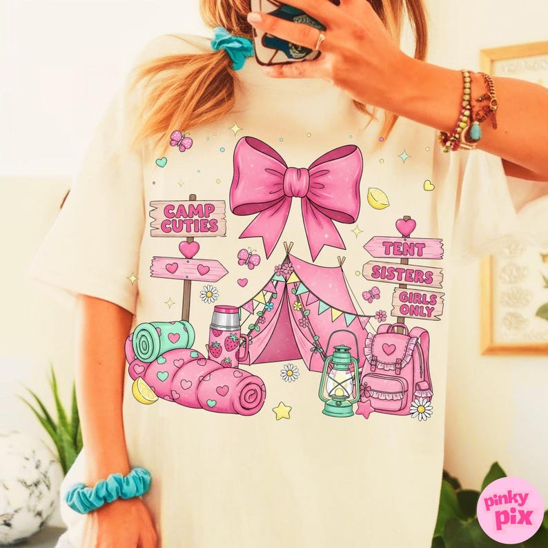 May include: Cream t-shirt with a pink camping design. The design includes a tent, bow, and signs reading "Camp Cuties" and "Tent Sisters Girls Only." Hearts, butterflies, and a backpack are also featured.