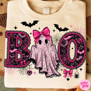 Puede incluir: Sudadera color crema con un dise&ntilde;o tem&aacute;tico de Halloween. La palabra "BOO" est&aacute; escrita en letras rosas con detalles de encaje negro y un lazo rosa. Un fantasma rosa con un lazo est&aacute; en el centro, rodeado de murci&eacute;lagos, telara&ntilde;as y una ara&ntilde;a.