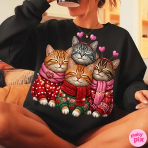 May include: Black sweatshirt featuring a graphic of four smiling cats. The cats are wearing sweaters and scarves, with heart-shaped headbands. The sweaters are red and green with floral and heart patterns. The cats are various shades of orange, gray, and brown.