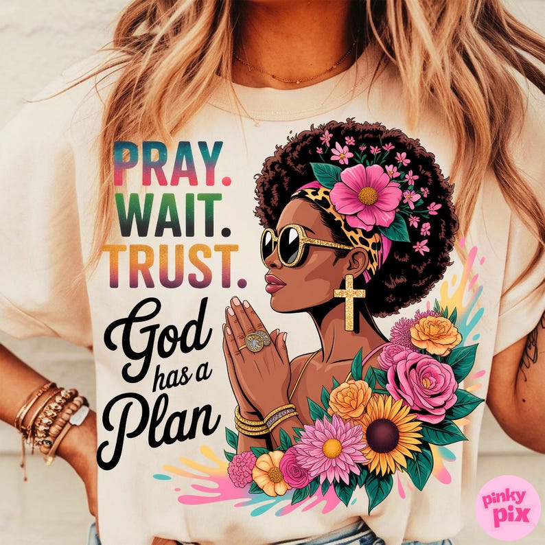 May include: Cream-colored t-shirt with the text "PRAY. WAIT. TRUST. God has a Plan." Features a graphic of a woman with sunglasses, a cross earring, and floral accents. The design includes colorful text and a floral arrangement.