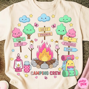 May include: A light beige t-shirt featuring a colorful camping-themed graphic. The design includes a campfire, smiling clouds, trees, and a backpack. Text includes "CAMPING CREW", "CAMP FUN", "S'MORES TIME", and "BEST FRIENDS".