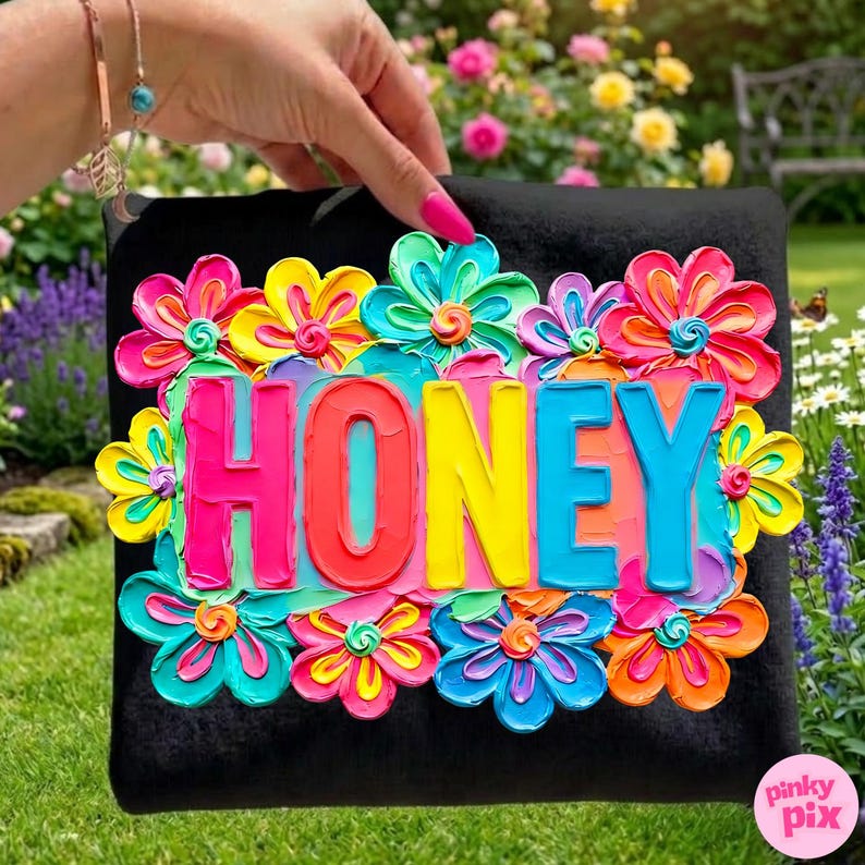 May include: Black decorative pillow with the word "HONEY" in vibrant, colorful letters. The word is surrounded by bright, textured flowers in shades of pink, yellow, blue, and orange. The pillow is held by a person.