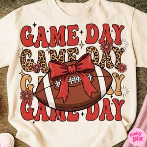 May include: Cream-colored t-shirt with a football graphic and the words "GAME DAY" in red, leopard print, and orange. A brown football with a red bow is in the center, with floral accents.