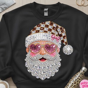 May include: A black crewneck sweatshirt featuring a graphic of Santa Claus wearing heart-shaped pink glasses. The design is composed of a mosaic of sparkling crystals in shades of brown, white, pink, and clear, forming a festive holiday pattern.