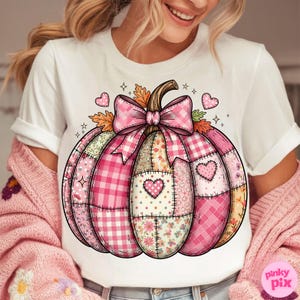 May include: White t-shirt featuring a pink patchwork pumpkin design. The pumpkin has a brown stem, a pink gingham bow, and heart accents. The design incorporates various pink patterns and floral elements.