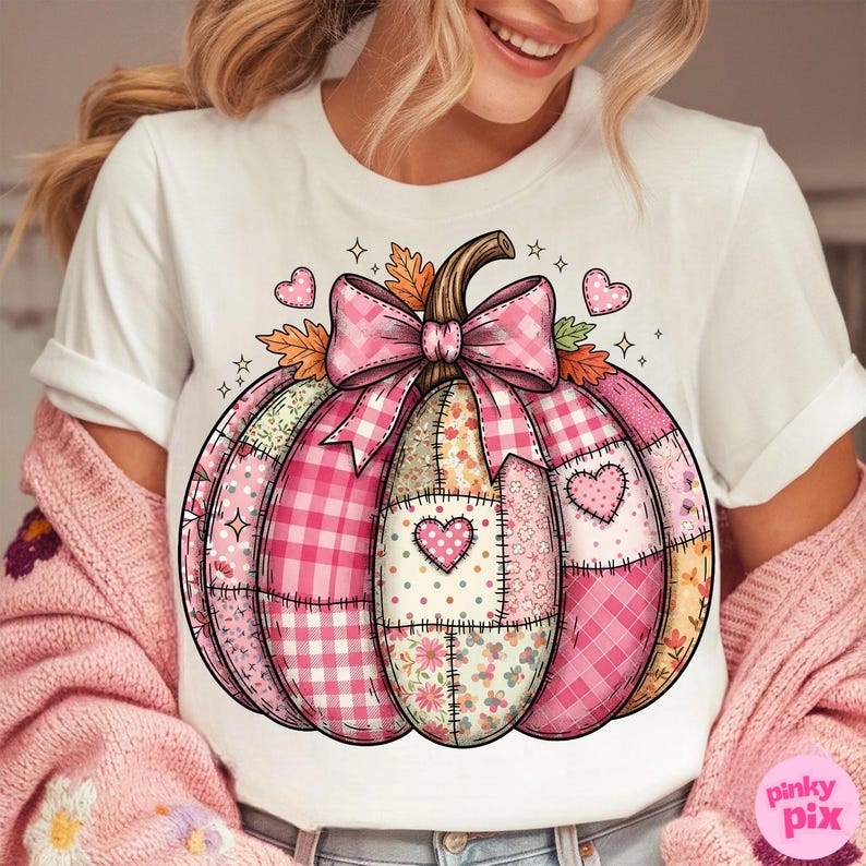 May include: White t-shirt featuring a pink patchwork pumpkin design. The pumpkin has a brown stem, a pink gingham bow, and heart accents. The design incorporates various pink patterns and floral elements.