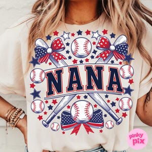 May include: A beige t-shirt with a graphic design featuring two crossed baseball bats, baseballs, red, white, and blue bows, and the word "NANA" in navy blue letters.