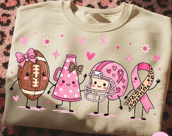 Könnte beinhalten: Ein beigefarbenes Sweatshirt mit Rundhalsausschnitt und einem Grafikdruck von vier anthropomorphen Figuren: ein brauner Football mit rosa Schleife, ein rosa Megafon mit Punkten, ein rosa Football-Helm und eine rosa Schleife mit Leopardenmuster.
