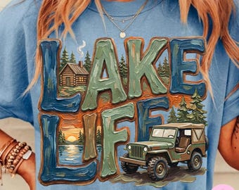Peut inclure: T-shirt bleu clair avec l'inscription "LAKE LIFE" dans un design texturé multicolore. Le motif comprend une cabane, un coucher de soleil sur un lac et une jeep verte.
